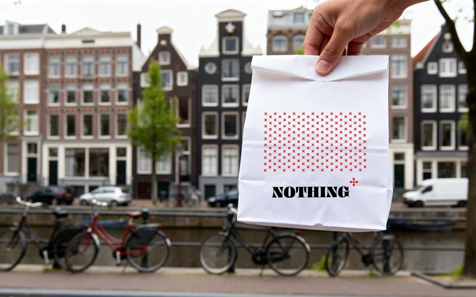 Un sacchetto di carta bianco con un rettangolo di croci rosse e il logo «NOTHING», sollevato in una strada lungo un canale.