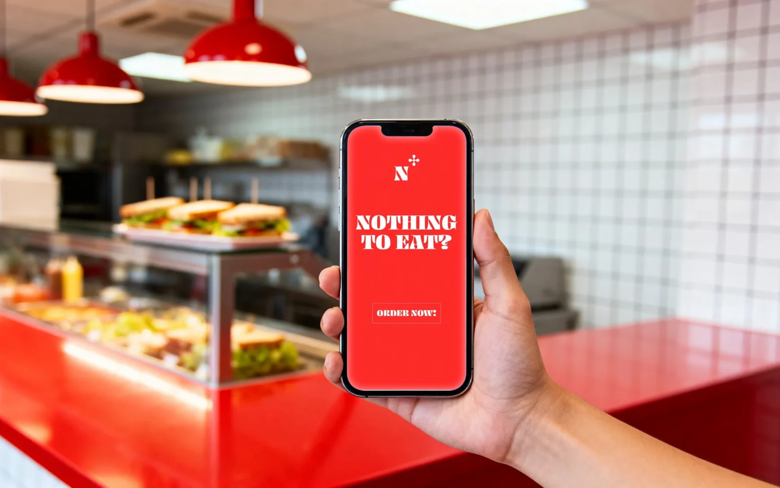 Una mano tiene uno smartphone nel locale; lo schermo rosso dice «NOTHING TO EAT?» con un pulsante «ORDER NOW!».