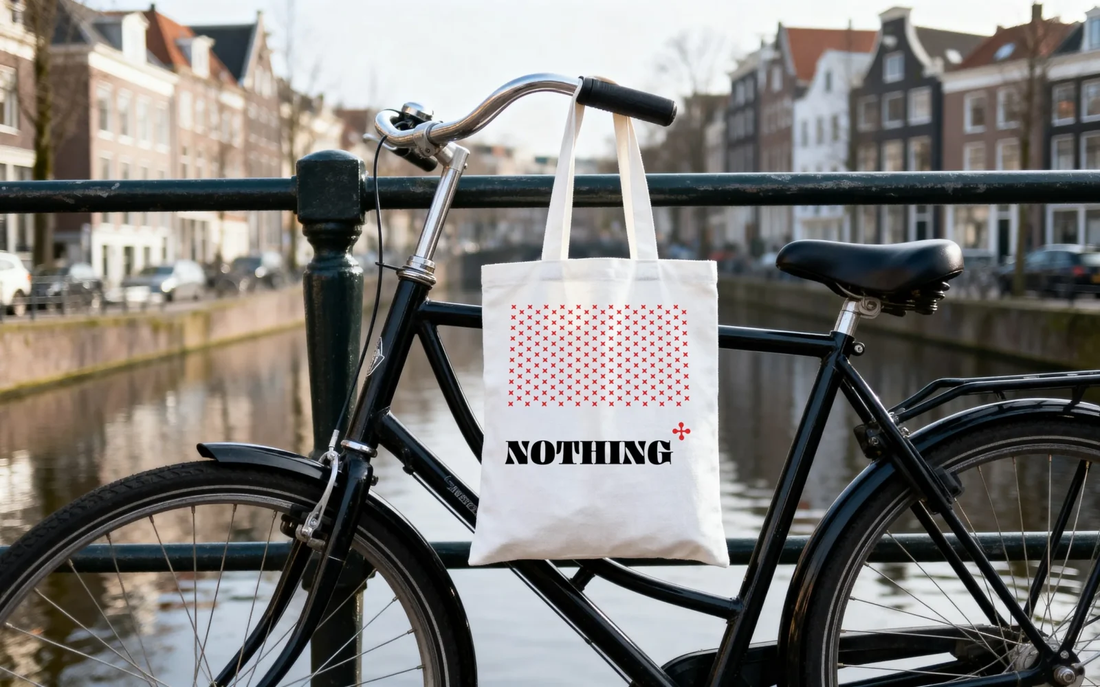 Una tote bag bianca con un rettangolo di croci rosse e il logo, appesa a una bicicletta nera lungo un canale.