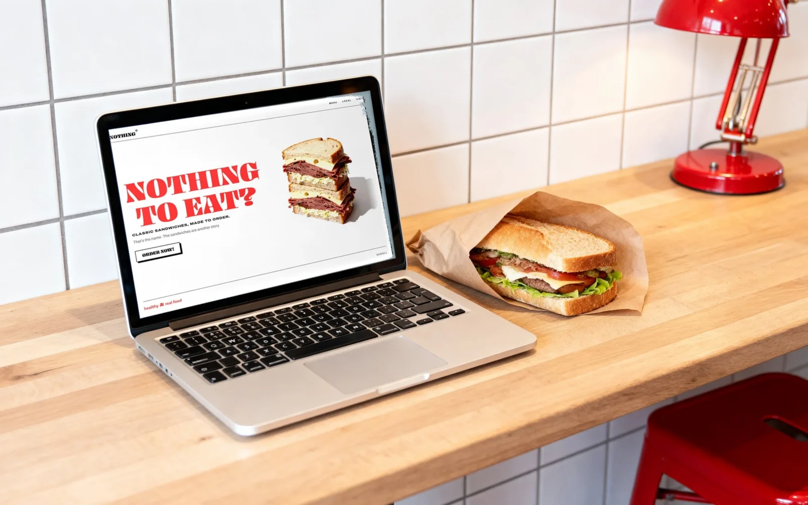 Un portatile su un bancone di legno davanti a piastrelle bianche, una lampada rossa sopra e un panino incartato accanto; lo schermo mostra la prima schermata del sito di Nothing To Eat con il lettering rosso «NOTHING TO EAT?».