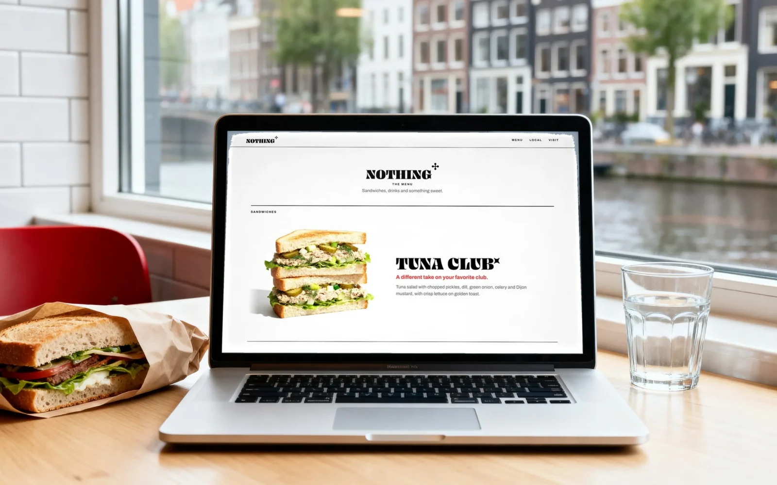 Un portatile su un tavolo a una finestra su un canale di Amsterdam, con un panino e un bicchiere d’acqua accanto; lo schermo mostra il menu del sito di Nothing To Eat con il Tuna Club.
