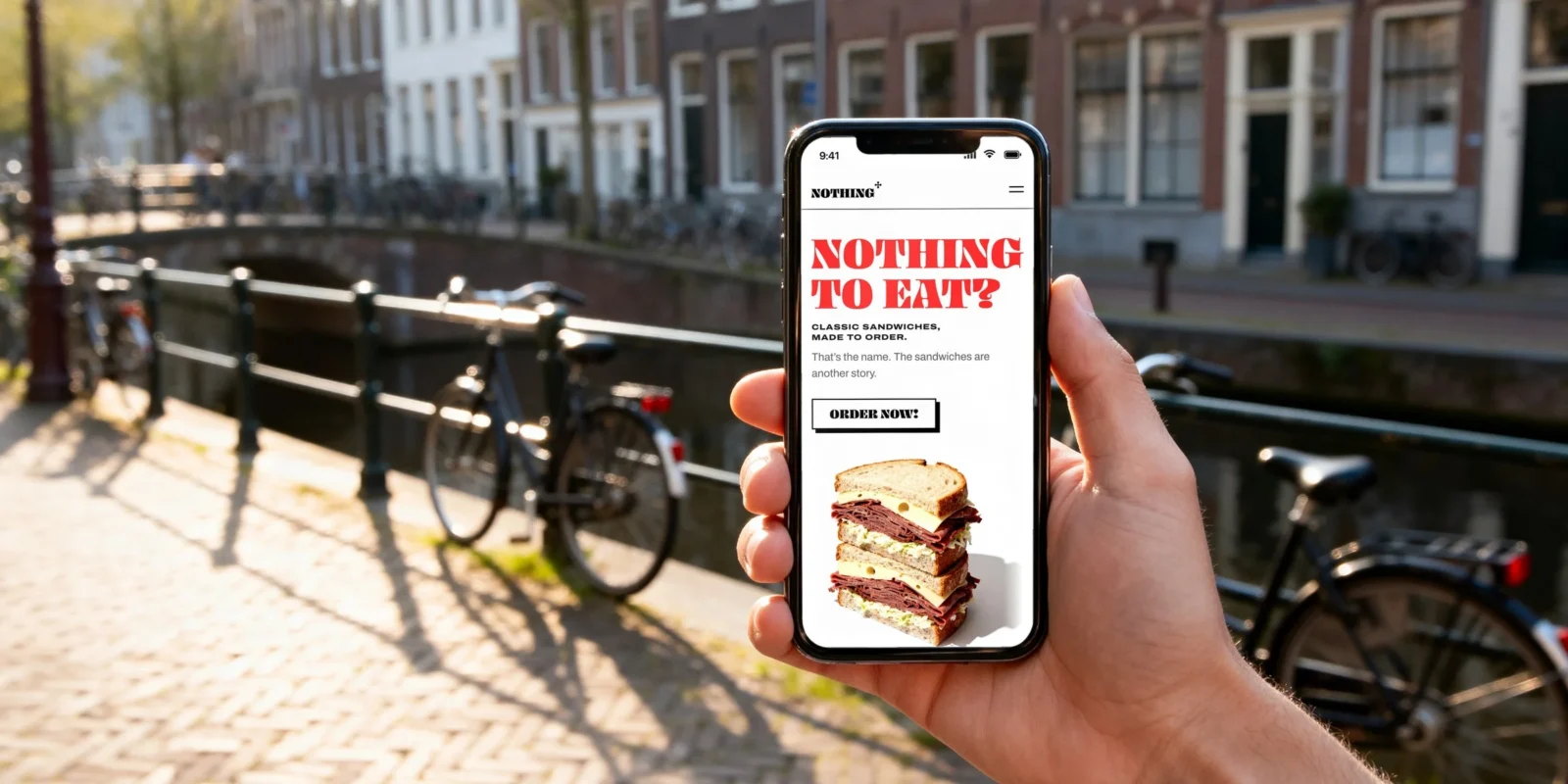 Una mano tiene uno smartphone lungo un canale di Amsterdam, con le biciclette contro la ringhiera alle spalle; lo schermo mostra il sito di Nothing To Eat: il lettering rosso «NOTHING TO EAT?», un pulsante «ORDER NOW!» e un panino al pastrami.