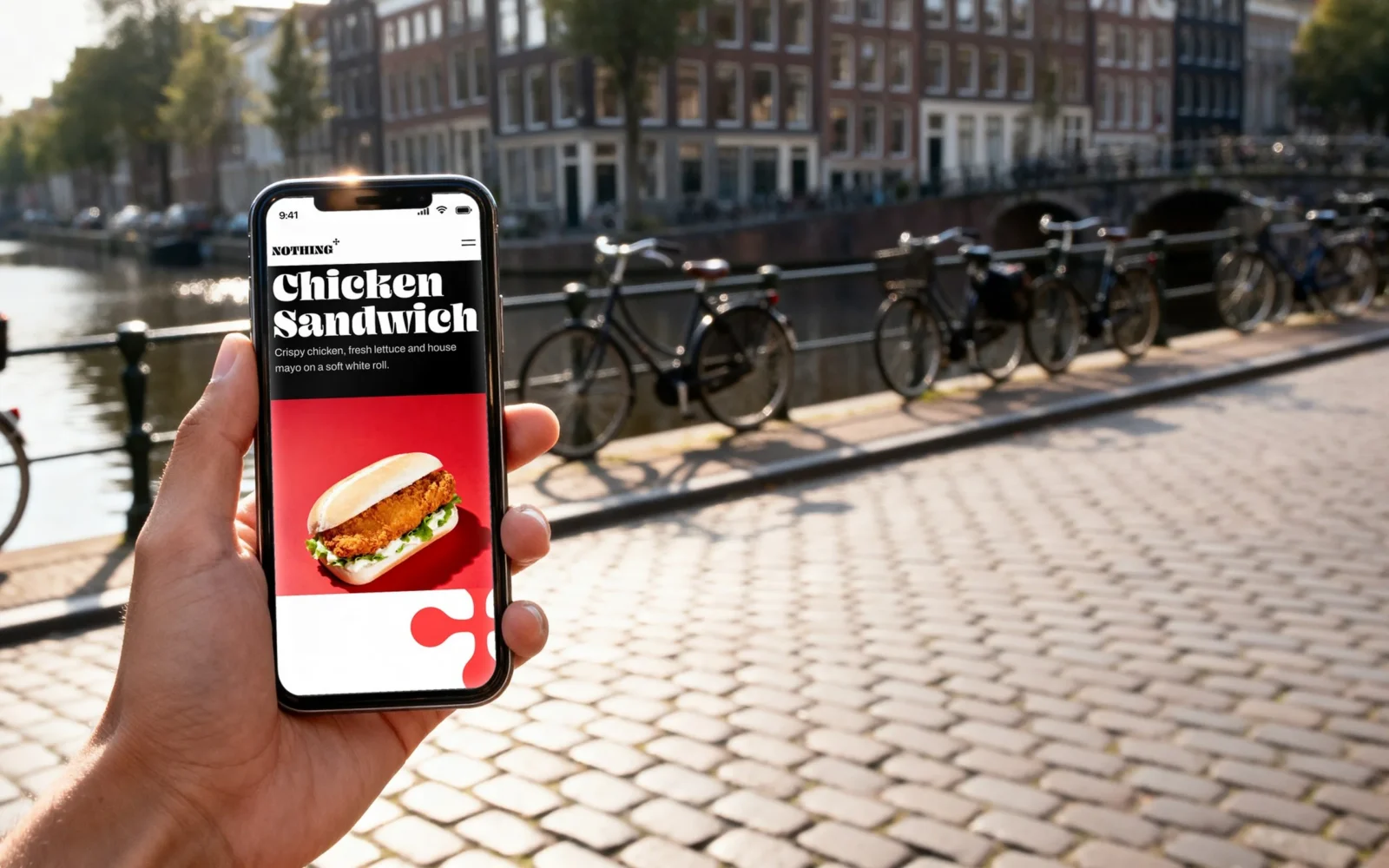 Una mano tiene uno smartphone sopra il selciato di una strada lungo un canale; lo schermo mostra la fascia nera e rossa del Chicken Sandwich.