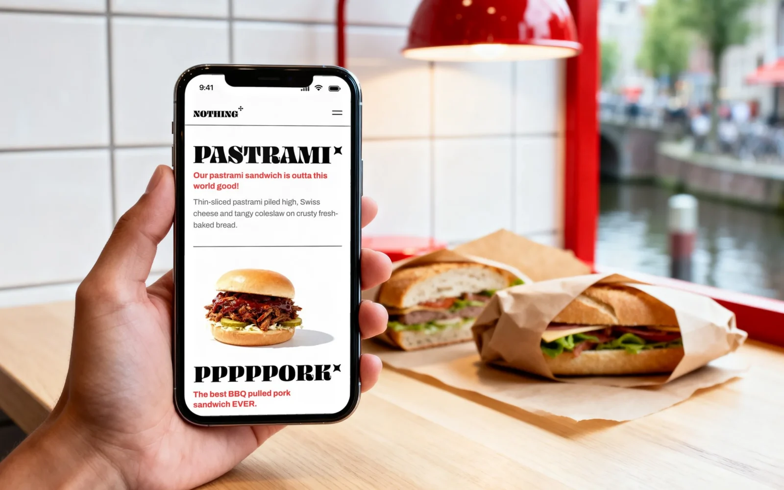 Una mano tiene uno smartphone sopra un bancone con panini incartati; lo schermo mostra il Pastrami e il Pppppork del menu di Nothing To Eat.