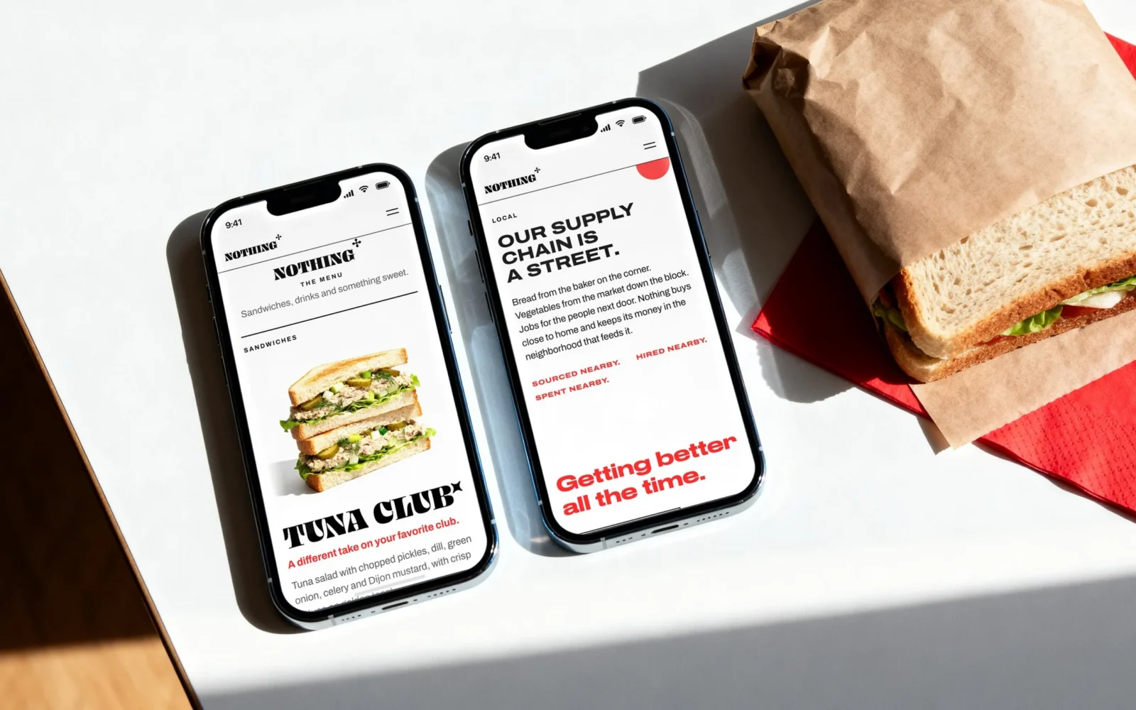 Due smartphone su un tavolo bianco accanto a un panino incartato e a un tovagliolo rosso; uno mostra il menu con il Tuna Club, l’altro la sezione «OUR SUPPLY CHAIN IS A STREET.»